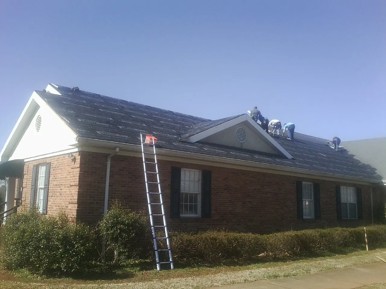 Crew tearing shingles off a brick ranch roof with a ladder set — Metal Roof Repair work by our crew in Efland