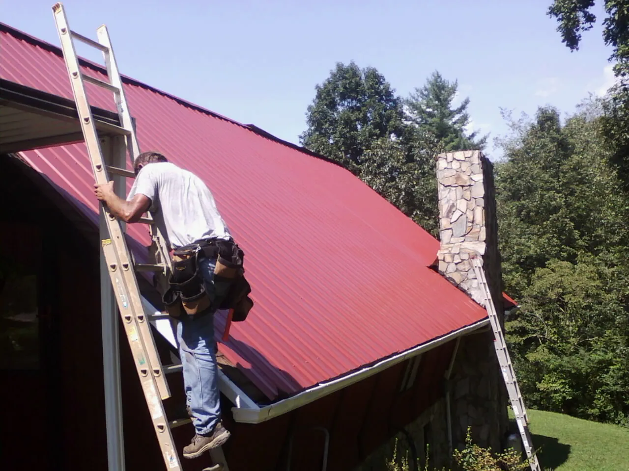Roofer on a ladder beside a red metal roof and stone chimney — Soffit & Fascia Repair by Roofing Brothers in Efland